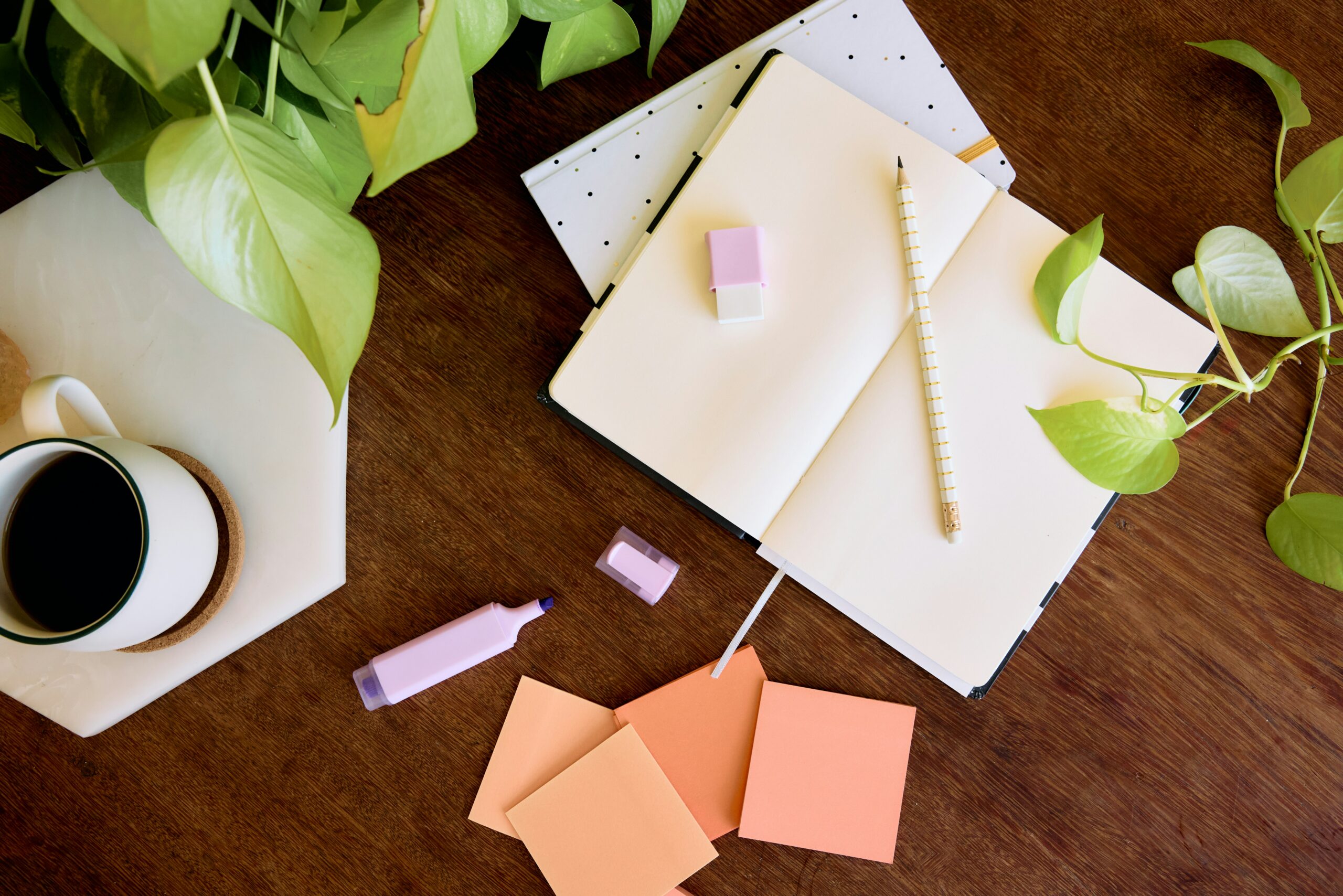 An open notebook with a striped pencil, sticky notes, a highlighter, and a cup of coffee on a wooden desk surrounded by green plants.