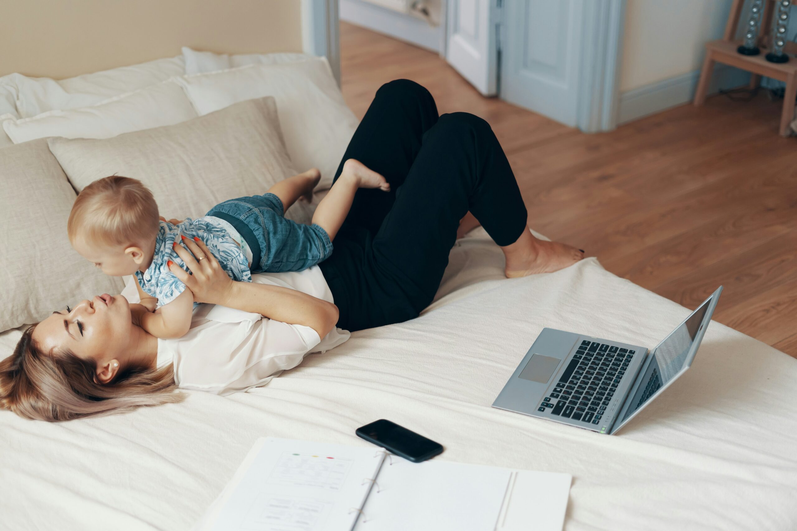 A woman lying on a bed playing with her baby, with a laptop and documents nearby.